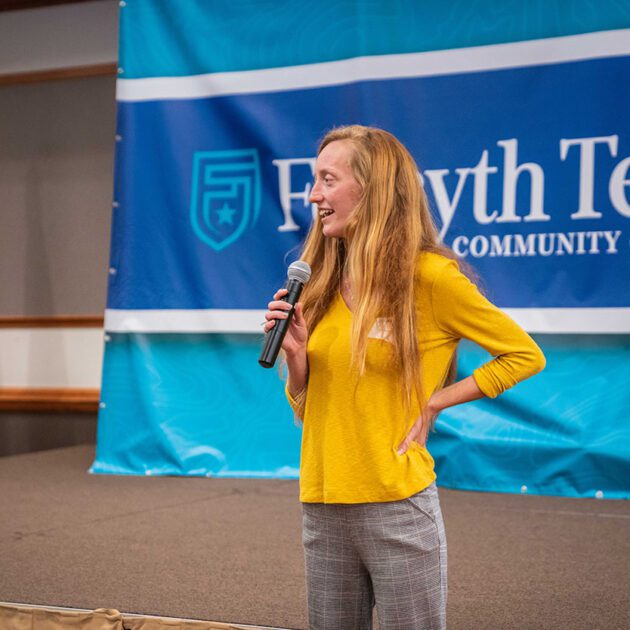 image woman wearing yellow shirt, long blonde hair speaking into microphone with forsyth tech banner behind