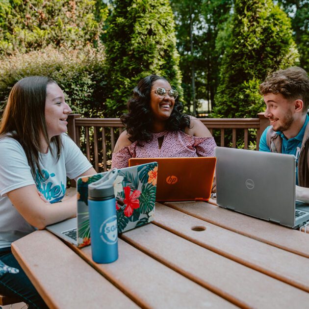 students sitting at a table outside working on laptops laughing