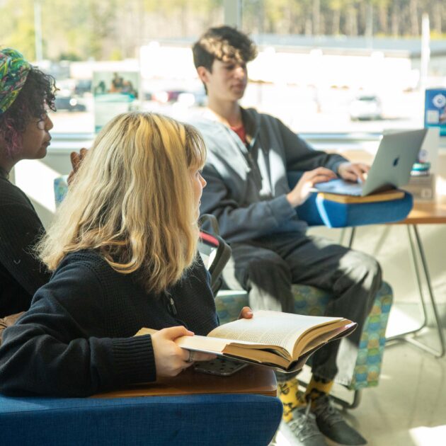 3 students sitting indoors beside a large window, one student has a book in hand and one is using a laptop