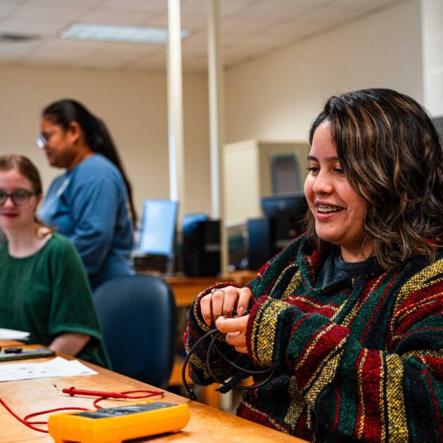 Image of a female student sitting in front of a machine working with wires. She is wearing a colorful sweater and smiling. There are two other female students in the background. They are inside of a computer lab.