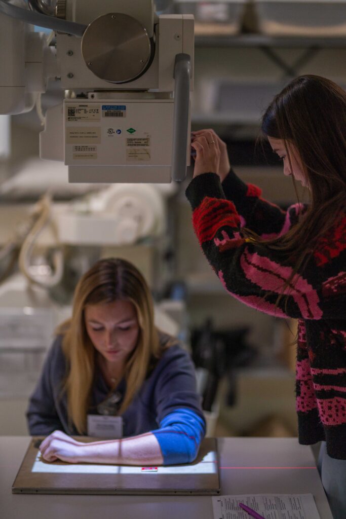 A woman sits with her arm under a radiography machine while another woman positions the machine