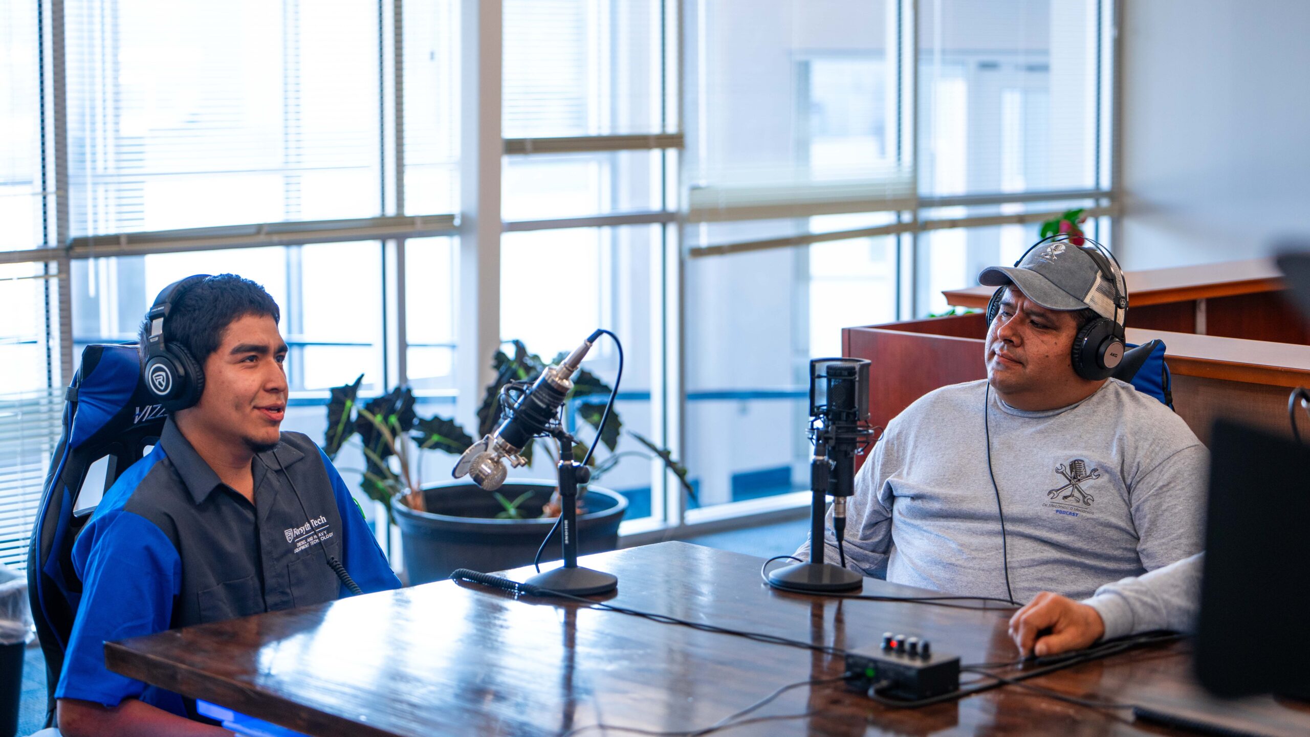 A young Latine man wearing a blue and grey button-up wears headphones and speaks to a man, also wearing headphones a baseball cap and grey long-sleeved tee shirt, at a table. They both have large microphones on the table in front of them.
