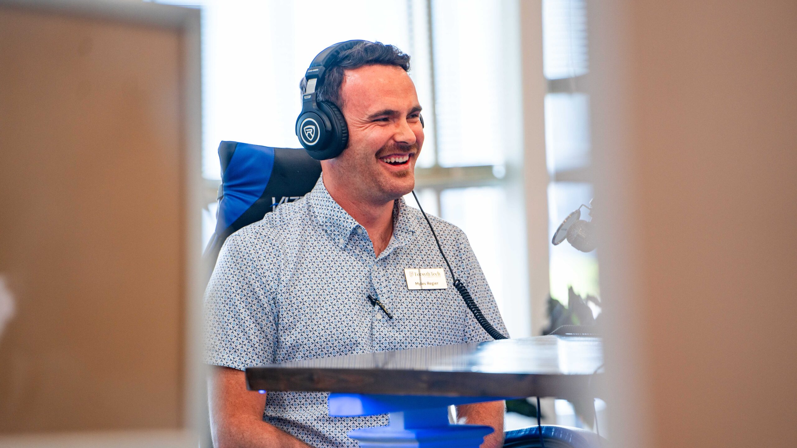 A white man with dark hair and a mustache wears headphones and light blue button-up shirt. He is smiling.