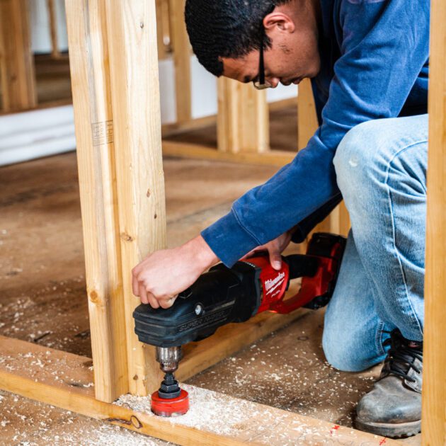A man wearing a blue shirt, jeans and boots is using a power tool on wood.