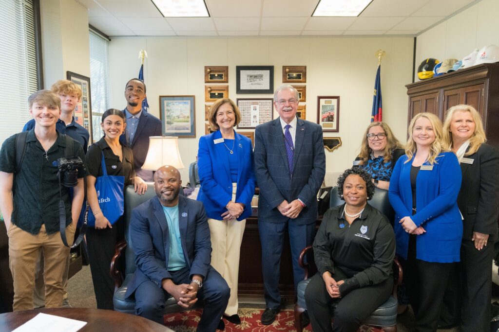 Un grupo de unas 12 personas, algunas sentadas, otras de pie, todas de diferentes edades y razas, se encuentran en una habitación interior que parece una oficina. Todos visten profesionalmente y sonríen a la cámara.