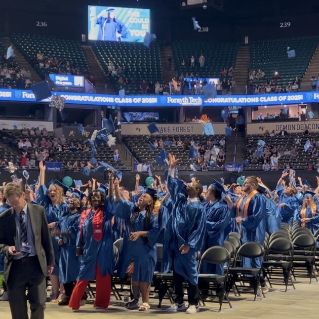 Thousands of people are in a coliseum indoors. In the foreground are graduates wearing royal blue gowns, smiling and tossing their royal blue graduation caps in the air.