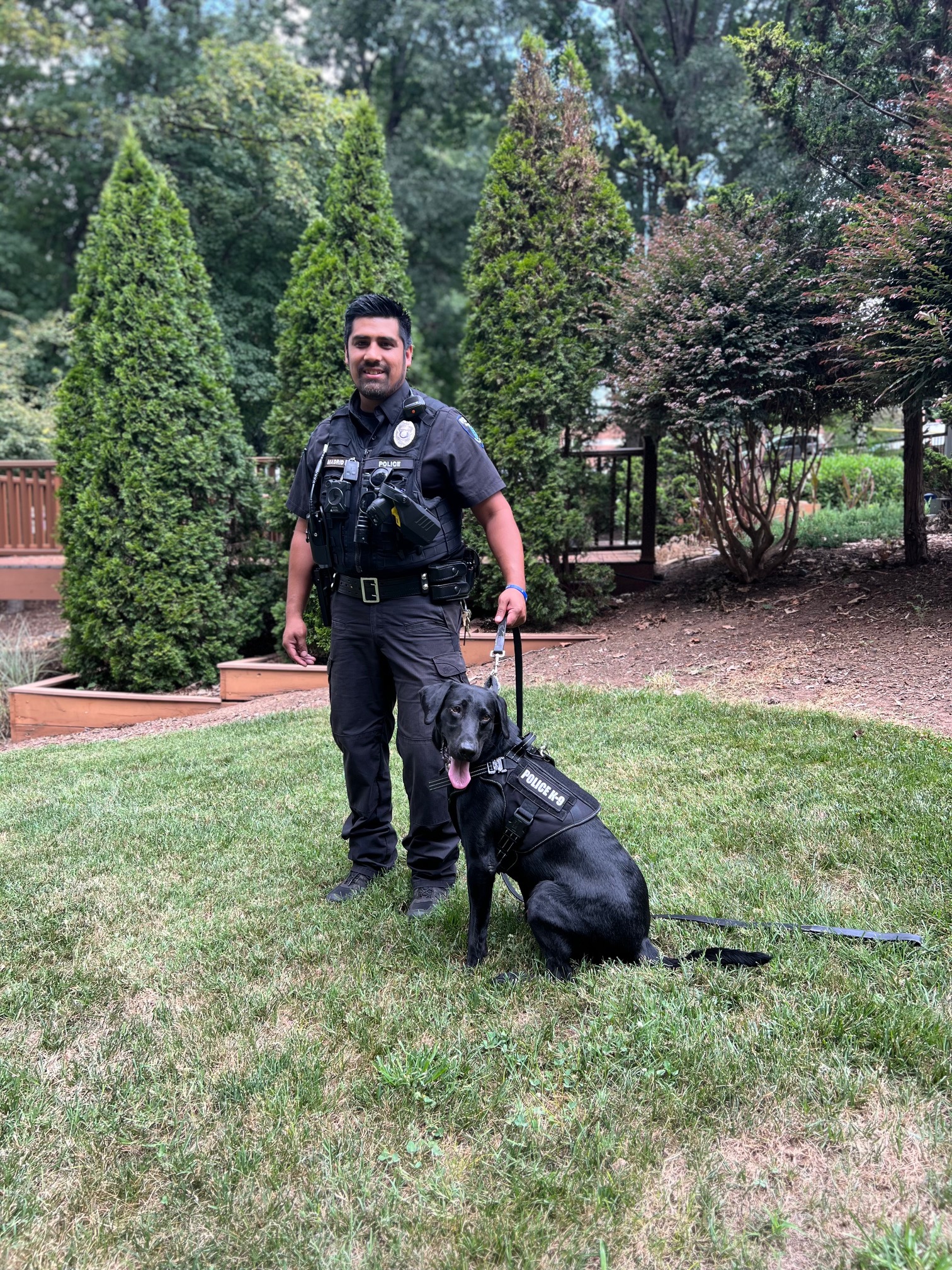 A man in a black police uniform with short, dark hair and facial hair stands outside, smiling, while holding a medium-sized black dog on a leash. The dog wears a vest that says "K-9 Officer)