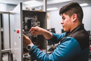Un estudiante trabaja en un panel de control eléctrico, utilizando un destornillador para ajustar el cableado y los componentes durante un laboratorio práctico de mecatrónica en Forsyth Tech.