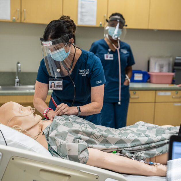 A female medical student is working on a mannequin in a classroom setting, another female medical student stands behind her.