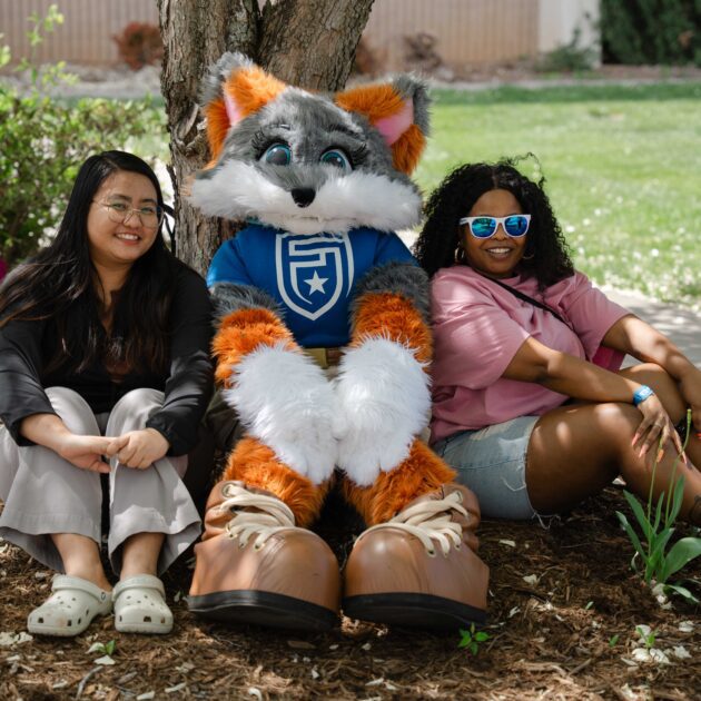 Two students are sitting beside Blaze the mascot. They are outside sitting on the ground in front of a tree. They are all smiling at the camera.