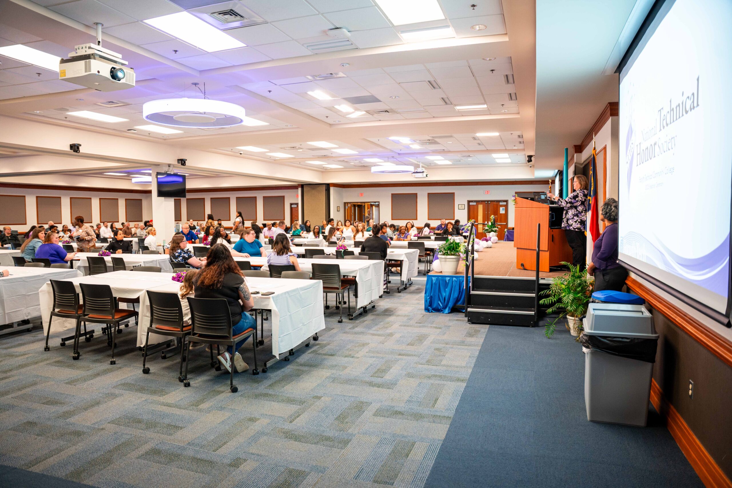 Several people sit at tables with white tablecloths inside of a conference room. They are all looking towards the stage.