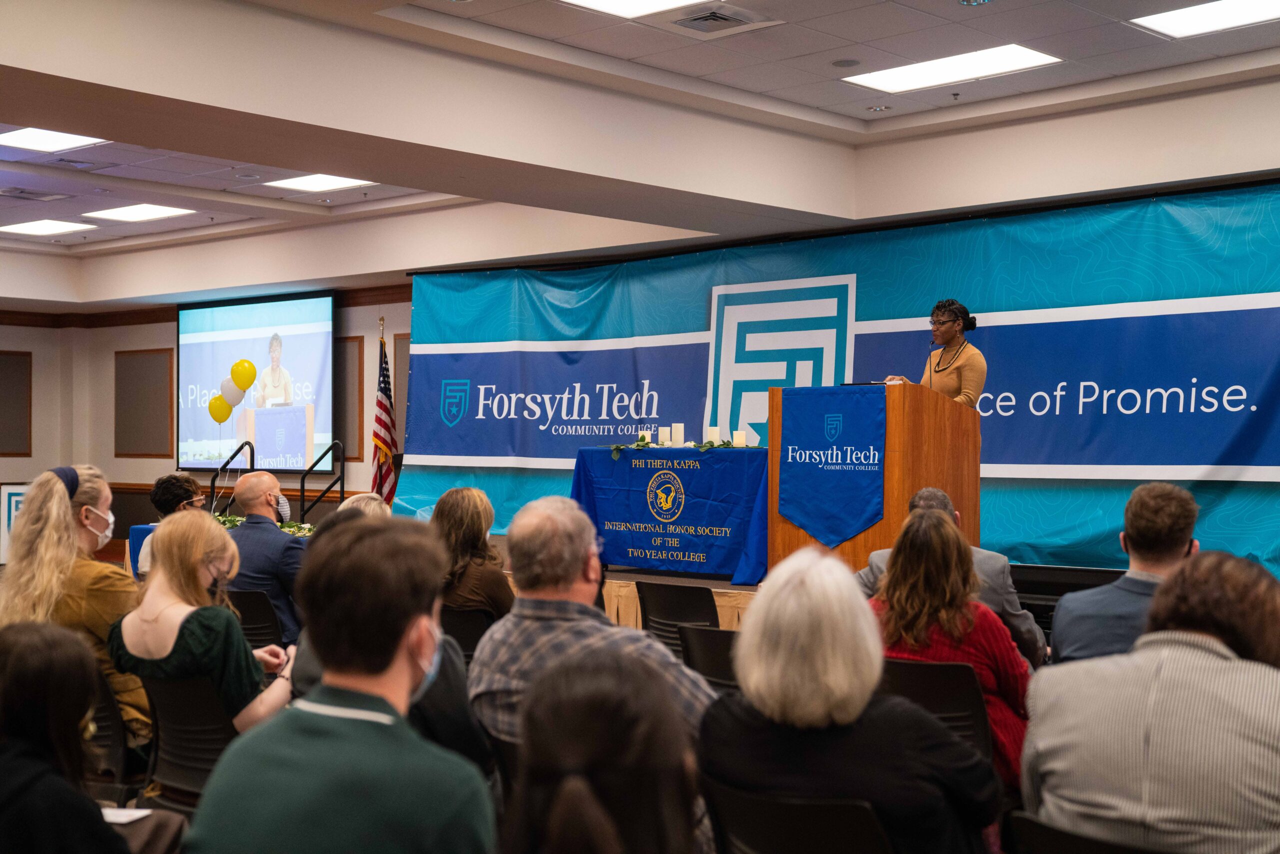 A woman wearing a yellow shirt is on stage speaking at a podium, in the forefront many people are sitting down and looking at her.