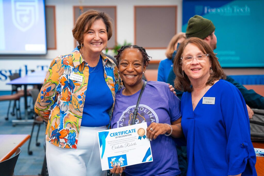 Two women stand on both sides of a student holding a certificate. All three are smiling at the camera.