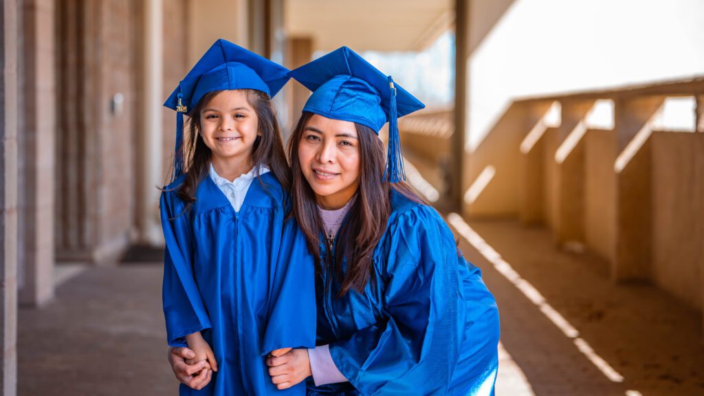 A student and her child, both wearing blue cap and gowns, smile at the camera. They are outside in warm lighting.