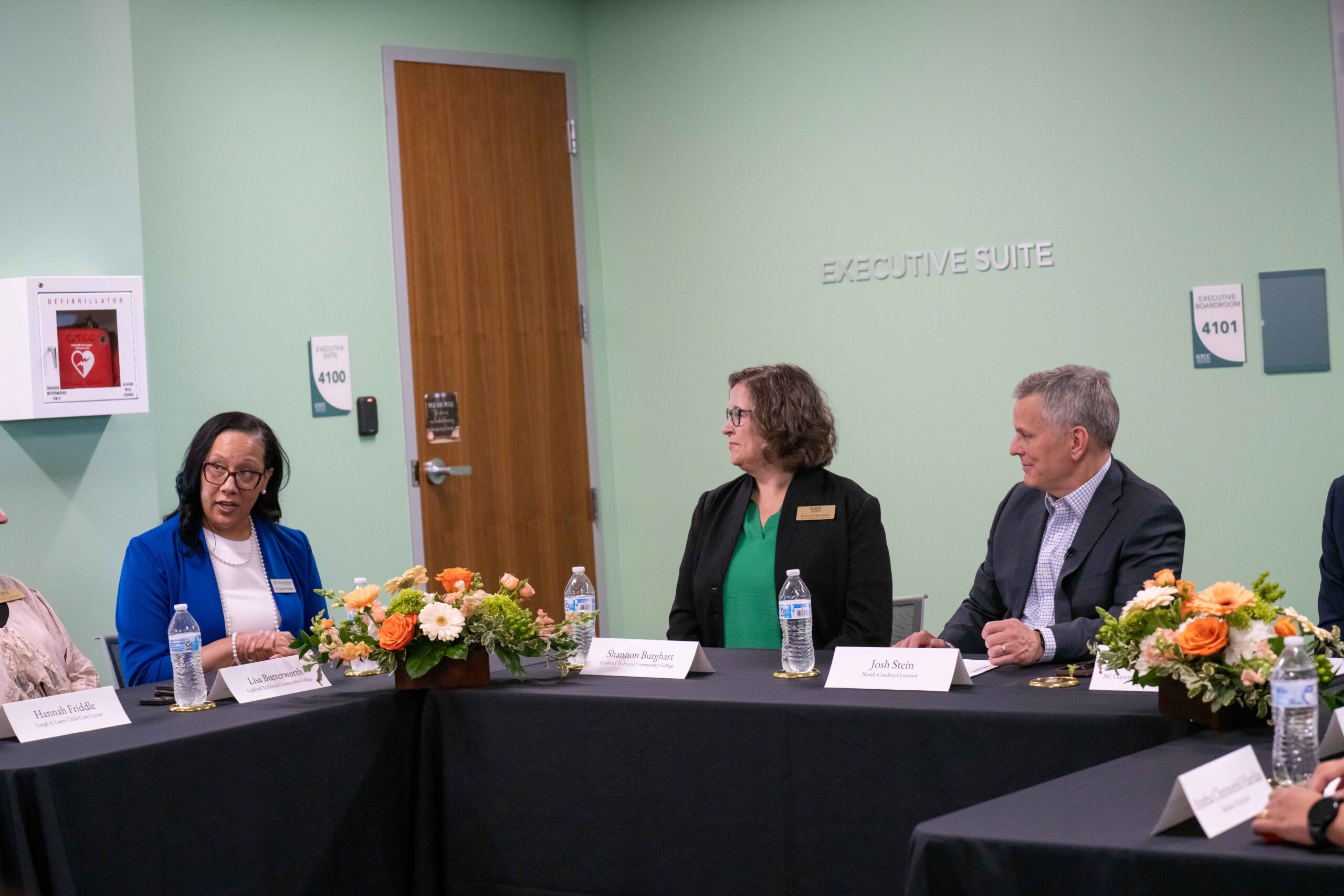 A group of professionals sits around a conference table in a room labeled “Executive Suite,” engaged in a discussion. Three individuals are in focus, with name placards, water bottles, and floral centerpieces on the table. One woman in a blue blazer appears to be speaking while others listen attentively.