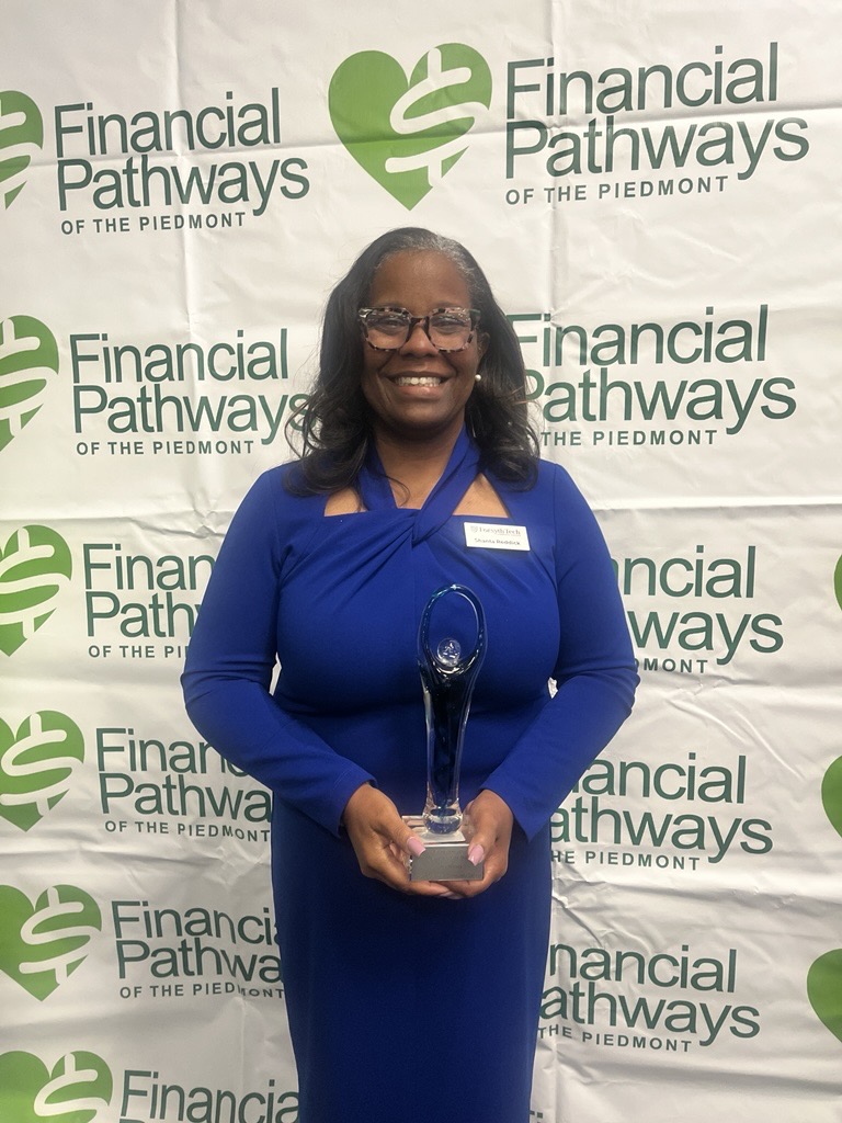 A woman with brown skin and black hair wears a royal blue dress. She is smiling at the camera and holding a clear award statue. She is standing in front of a backdrop that says "Financial Pathways of the Piedmont"